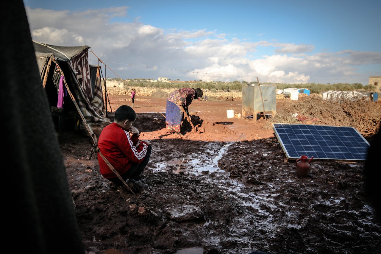 Services-01 A boy and woman in a muddy refugee camp in Idlib, Syria with tents and solar panel.